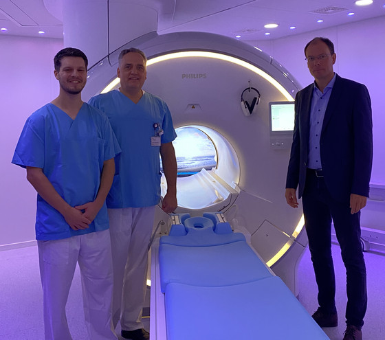 Richard Grundherr, Prof. Christoph Weber und Andreas Schneider stehen in einem dunkellila illuminierten Raum vor dem neuen MRT-System im Immanuel Albertinen Zentrum für Radiologie am Standort Ev. Amalie Sieveking Krankenhaus in Hamburg und blicken in die Kamera